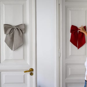 Può includere: Due fiocchi decorativi sono montati su porte bianche. Uno è a motivo a scacchi grigio e bianco, l'altro è rosso pieno. Una persona sta raggiungendo il fiocco rosso.