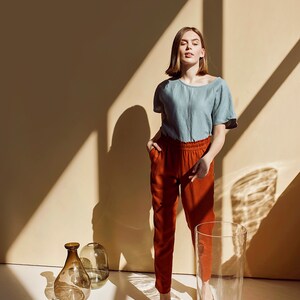 May include: A woman wearing a light blue linen shirt and rust-colored pants stands in a room with a beige wall and sunlight streaming through the window. She is surrounded by various glass vases and jars.