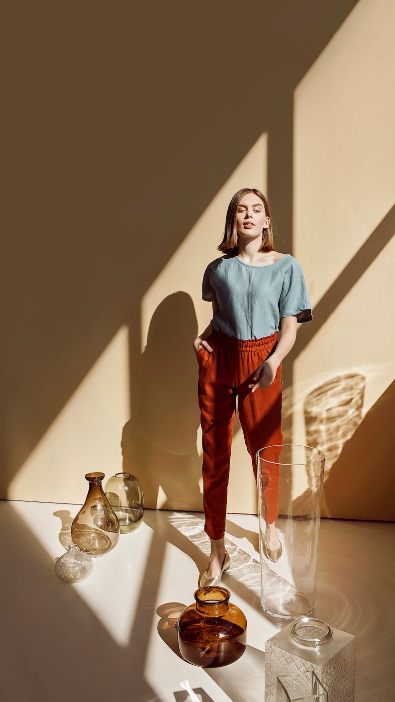 May include: A woman wearing a light blue linen shirt and rust-colored pants stands in a room with a beige wall and sunlight streaming through the window. She is surrounded by various glass vases and jars.