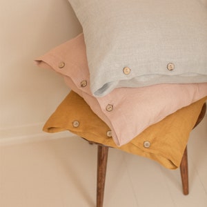 May include: Three linen pillowcases stacked on a wooden stool. The top pillowcase is light gray, the middle pillowcase is light pink, and the bottom pillowcase is mustard yellow. Each pillowcase has wooden buttons.
