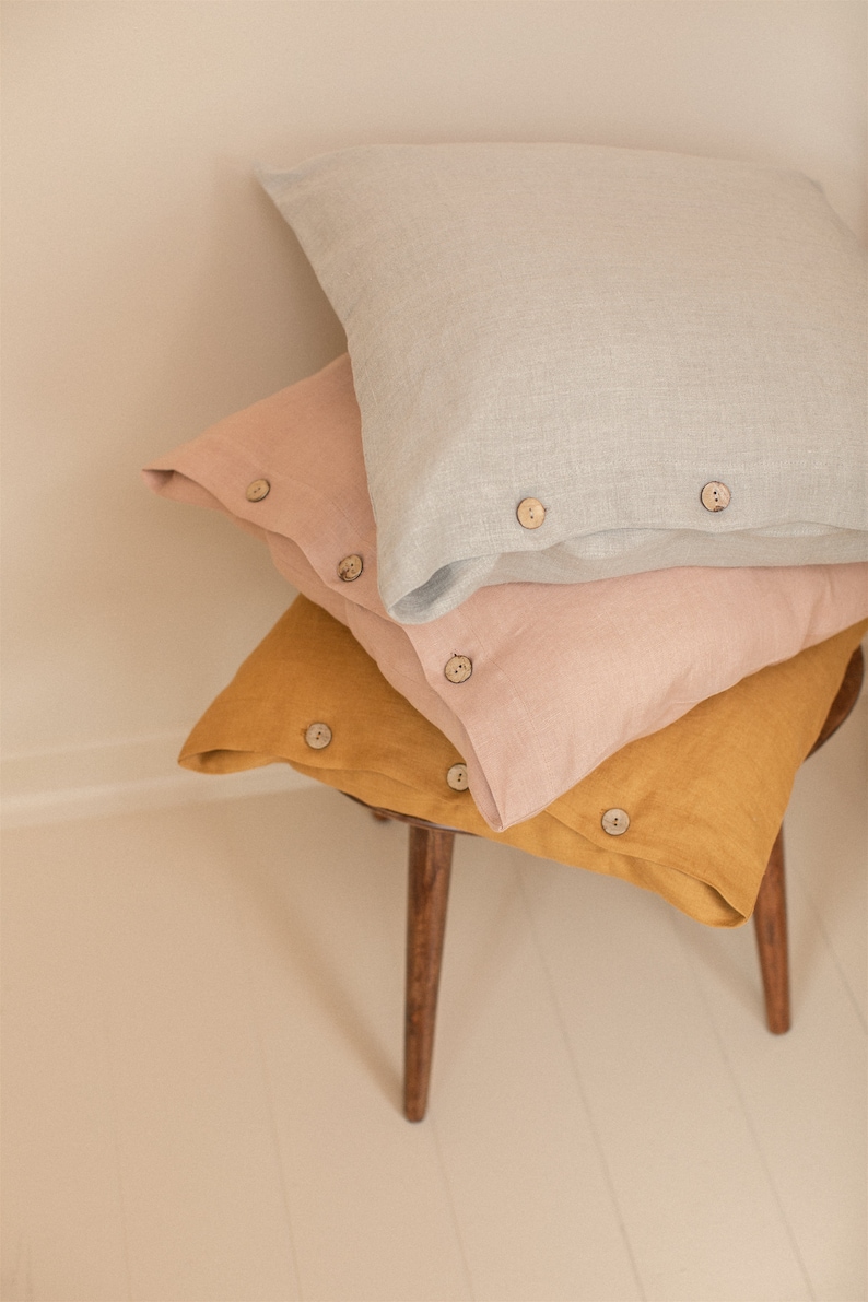 May include: Three linen pillowcases stacked on a wooden stool. The top pillowcase is light gray, the middle pillowcase is light pink, and the bottom pillowcase is mustard yellow. Each pillowcase has wooden buttons.