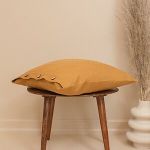 May include: A mustard yellow linen pillow with wooden buttons sits on a wooden stool with three legs. The stool is on a light beige floor.