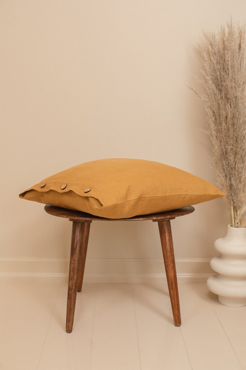 May include: A mustard yellow linen pillow with wooden buttons sits on a wooden stool with three legs. The stool is on a light beige floor.