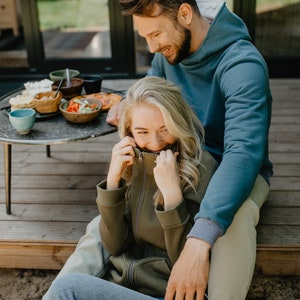 May include: A couple is seated on a wooden deck, enjoying a meal. The woman wears an olive green zip-up hoodie, while the man wears a blue hoodie. A small, round table holds bowls of food and drinks. The scene suggests a relaxed outdoor gathering.