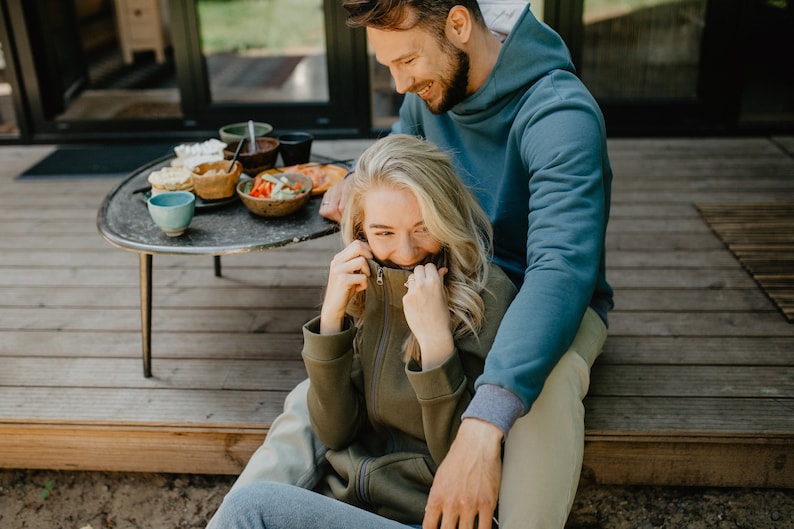 May include: A couple is seated on a wooden deck, enjoying a meal. The woman wears an olive green zip-up hoodie, while the man wears a blue hoodie. A small, round table holds bowls of food and drinks. The scene suggests a relaxed outdoor gathering.