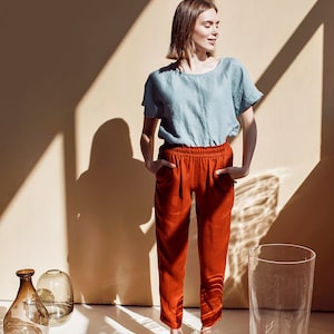May include: A woman wearing a light blue linen top and red pants stands in a room with sunlight streaming through the windows. She is smiling and looking to the side. There are several glass vases on the floor.