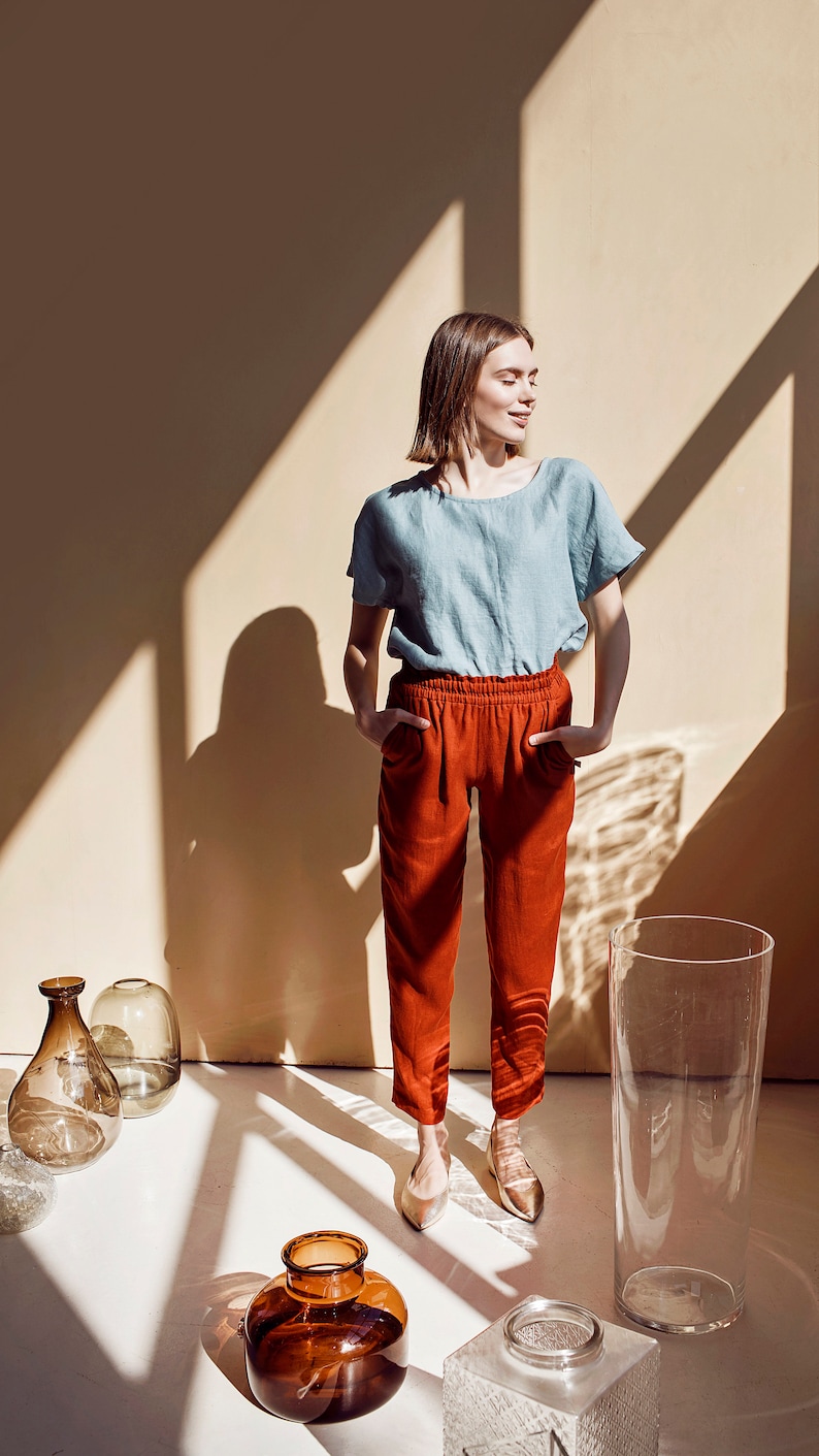 May include: A woman wearing a light blue linen top and red pants stands in a room with sunlight streaming through the windows. She is smiling and looking to the side. There are several glass vases on the floor.