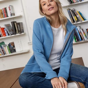May include: A light blue zip-up jacket with a gray interior lining. The jacket is worn over a white t-shirt and paired with blue jeans. The person is sitting on a brown surface in front of a bookshelf.