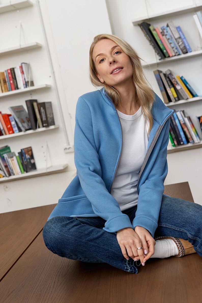 May include: A light blue zip-up jacket with a gray interior lining. The jacket is worn over a white t-shirt and paired with blue jeans. The person is sitting on a brown surface in front of a bookshelf.