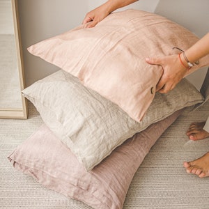 May include: Three linen pillow covers in shades of pink and beige. The top pillow cover is being held by a hand.