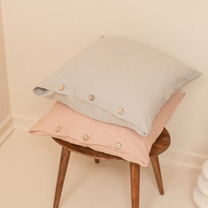 May include: Two linen pillowcases stacked on a wooden stool. The top pillowcase is light gray and the bottom pillowcase is a light peach color. Both pillowcases have wooden buttons.