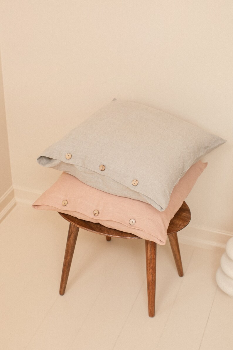 May include: Two linen pillowcases stacked on a wooden stool. The top pillowcase is light gray and the bottom pillowcase is a light peach color. Both pillowcases have wooden buttons.