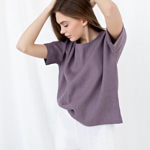 May include: A short-sleeved, lavender-colored linen top paired with white linen pants. The top has a round neckline and a relaxed fit. The model is wearing the outfit and has their hands in their hair.