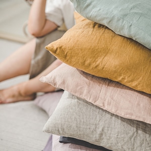 May include: A stack of five linen pillows in various colors, including light green, mustard yellow, light pink, gray, and purple. The pillows are arranged in a pyramid shape.