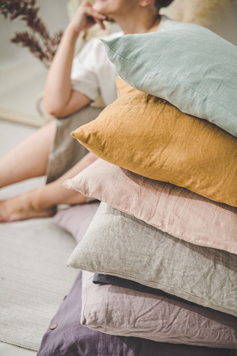 May include: A stack of five linen pillows in various colors, including light green, mustard yellow, light pink, gray, and purple. The pillows are arranged in a pyramid shape.