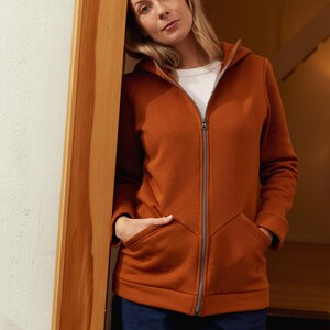 May include: A rust-colored zip-up hoodie with a hood and front pockets. The jacket is worn over a white t-shirt and paired with dark blue jeans. The zipper is silver-toned.