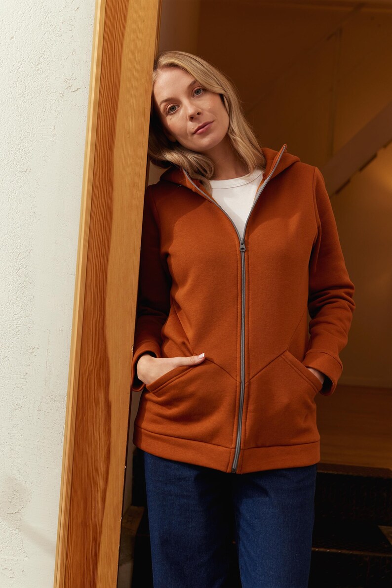 May include: A rust-colored zip-up hoodie with a hood and front pockets. The jacket is worn over a white t-shirt and paired with dark blue jeans. The zipper is silver-toned.