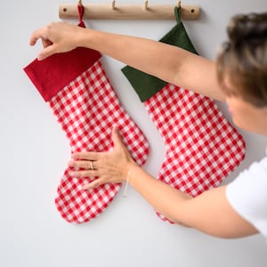 Könnte beinhalten: Zwei Weihnachtsstrümpfe mit rot-weißem Vichy-Muster und roten und grünen Manschetten hängen an einer Holzstange. Die Strümpfe werden an einer weißen Wand angebracht. Eine festliche Weihnachtsdekoration.