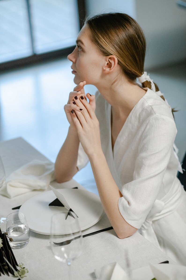 Bride in a white linen wrap dress sitting at a table, hand under her chin