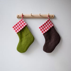 May include: Two Christmas stockings, one olive green and one dark brown, hang from a wooden rack. Each stocking has a red and white checkered cuff. The stockings are suspended against a white wall.