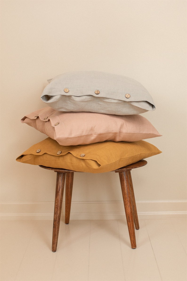 May include: Three linen pillow covers stacked on a wooden stool. The top pillow cover is light gray, the middle pillow cover is light pink, and the bottom pillow cover is mustard yellow. All three pillow covers have wooden buttons.
