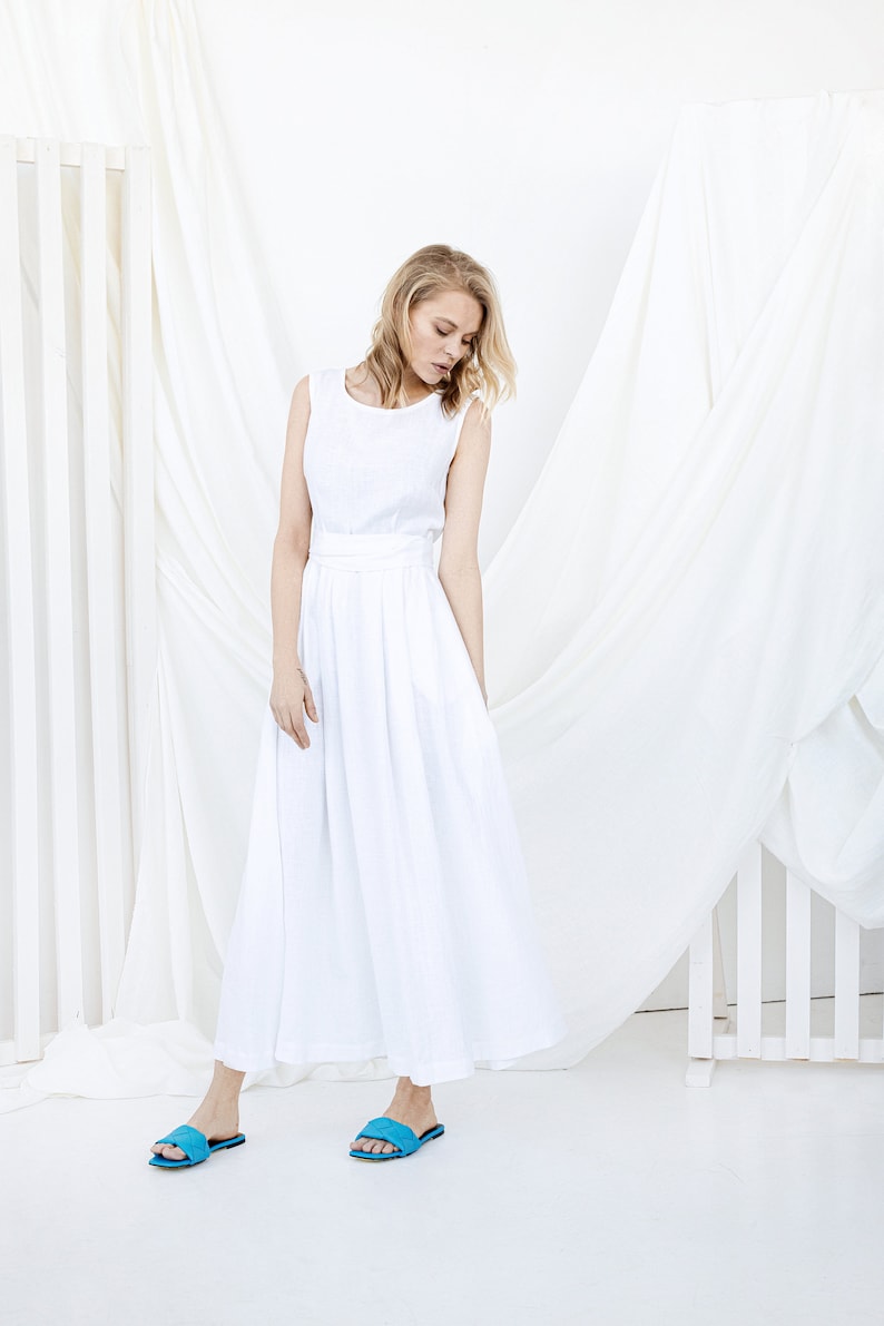 May include: A white linen dress with a sleeveless bodice and a flowing skirt. The dress has a belt at the waist and is worn with blue sandals.