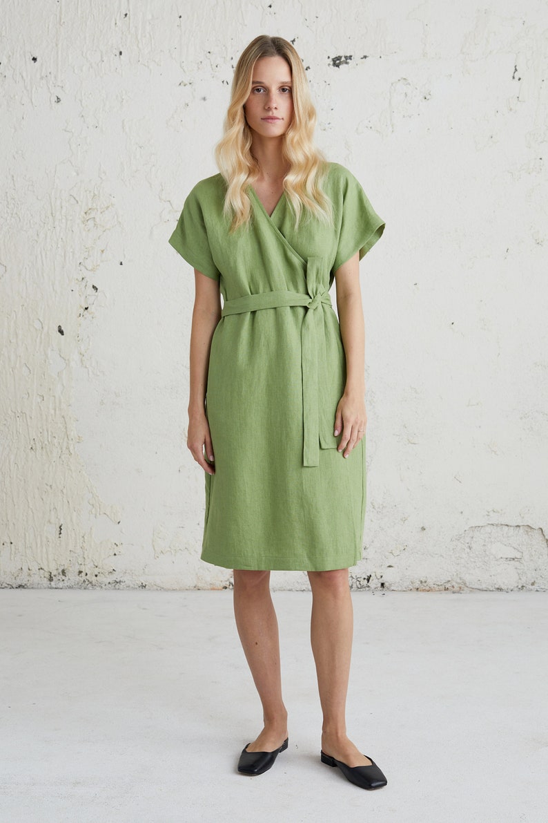 May include: A green linen wrap dress with short sleeves and a self-tie belt. The dress is knee-length and has a v-neckline.