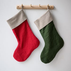 Könnte beinhalten: Zwei Weihnachtsstrümpfe, einer rot und einer grün, hängen an einer Holzstange vor einer weißen Wand. Jeder Strumpf hat eine neutralfarbene Manschette. Die Strümpfe sind aus einem strukturierten Stoff gefertigt.