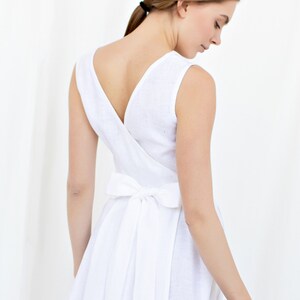 May include: A white linen dress with a V-neckline and a bow tie at the back. The dress is sleeveless and has a flowing skirt.
