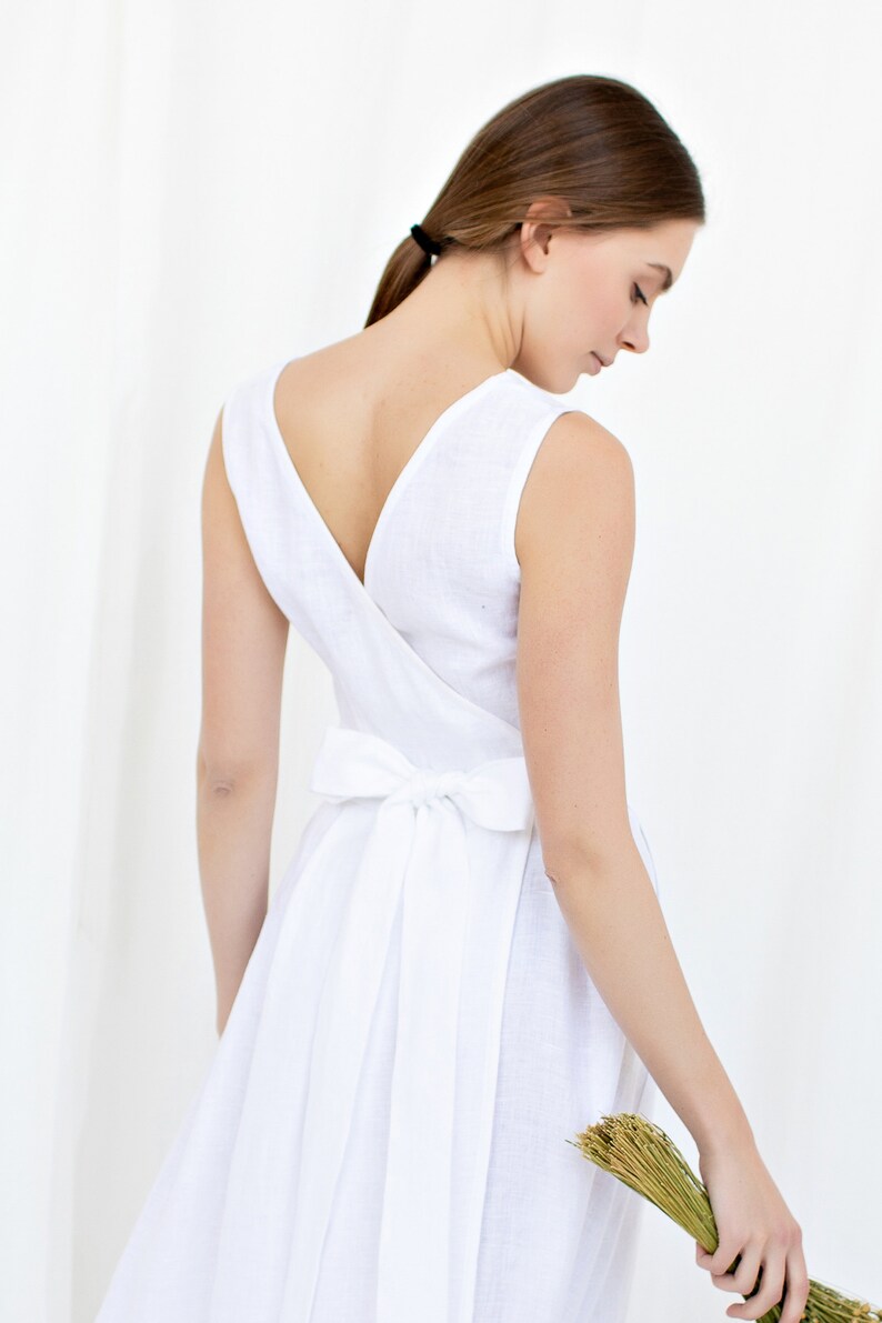 May include: A white linen dress with a V-neckline and a bow tie at the back. The dress is sleeveless and has a flowing skirt.
