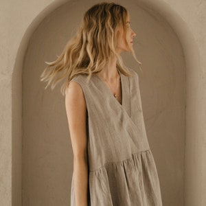 May include: A woman wearing a light brown linen dress with a V-neckline and a simple gold necklace. The dress has a flowing, relaxed fit and is sleeveless.