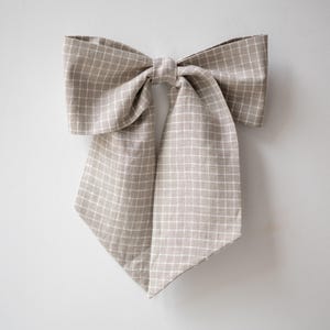 Può includere: Un grande fiocco beige con un motivo a griglia bianca. Il fiocco è legato al centro, con estremità lunghe e appuntite che pendono. Il tessuto sembra essere lino o un materiale simile. Il fiocco è un accessorio decorativo.