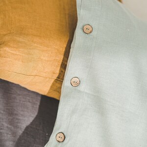 May include: Three pillowcases in different colors and textures. The top pillowcase is light green with three wooden buttons. The middle pillowcase is a dark gray linen. The bottom pillowcase is a mustard yellow linen.