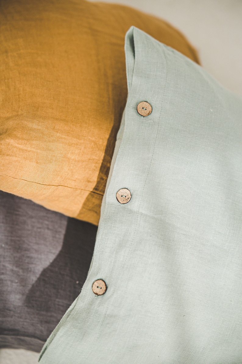 May include: Three pillowcases in different colors and textures. The top pillowcase is light green with three wooden buttons. The middle pillowcase is a dark gray linen. The bottom pillowcase is a mustard yellow linen.