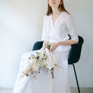 Bride in a white linen maxi wrap dress sitting on a chair with a bouquet