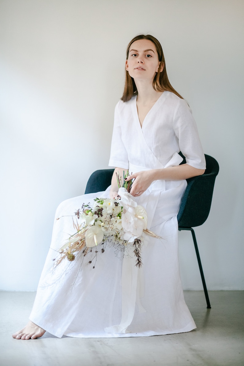 Bride in a white linen maxi wrap dress sitting on a chair with a bouquet