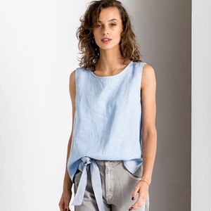 May include: A light blue sleeveless linen top with a tie detail at the waist. The top is paired with gray denim pants. The outfit is simple and elegant, suitable for casual wear.