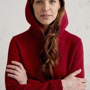 May include: A burgundy hooded sweatshirt with a full-length zipper. The sweatshirt has a high collar and a hood. The person in the image has their arms crossed. The background is a textured, off-white wall.