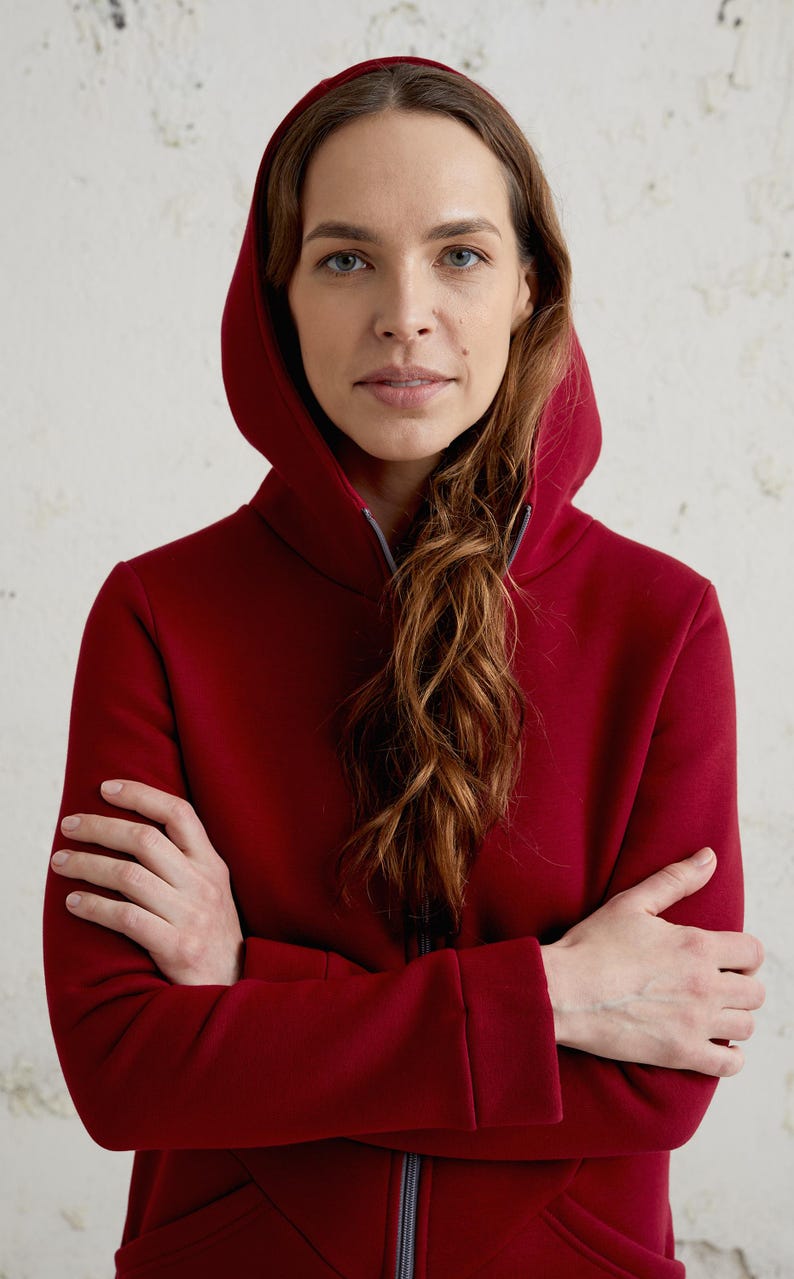 May include: A burgundy hooded sweatshirt with a full-length zipper. The sweatshirt has a high collar and a hood. The person in the image has their arms crossed. The background is a textured, off-white wall.