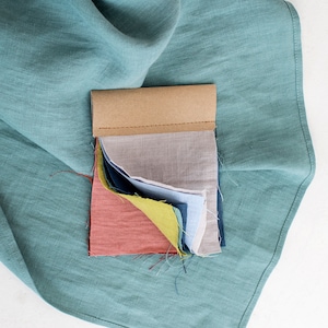 Può includere: Un libro di campioni di tessuto con una varietà di colori, tra cui beige, blu, verde, giallo e rosso. I campioni sono disposti in una pila e sono tenuti insieme da una fascia di carta marrone.