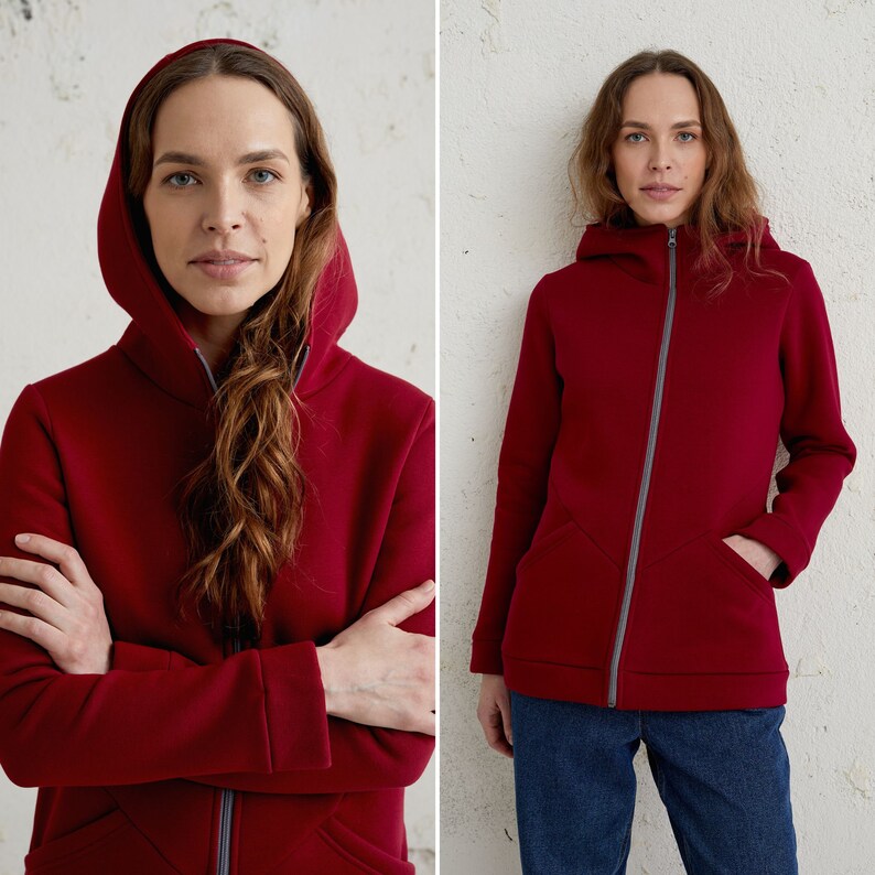 May include: A burgundy hooded sweatshirt with a full-length zipper and two front pockets. The hood is lined with gray fabric, and the zipper is also gray. The model is wearing blue jeans.