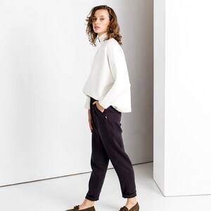 May include: A woman wearing a white turtleneck sweater and dark gray pants. She is standing in a white room and has her hand in her pocket. She is wearing dark green loafers.