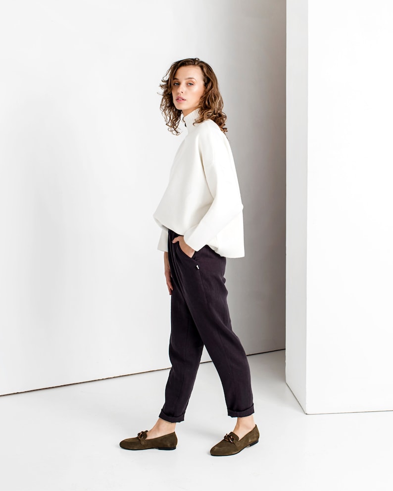 May include: A woman wearing a white turtleneck sweater and dark gray pants. She is standing in a white room and has her hand in her pocket. She is wearing dark green loafers.