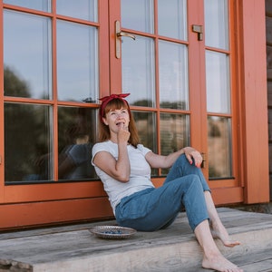 May include: A woman wearing a red headband sits on a wooden porch with a bowl of blueberries in front of her. She is eating a blueberry and looking up.