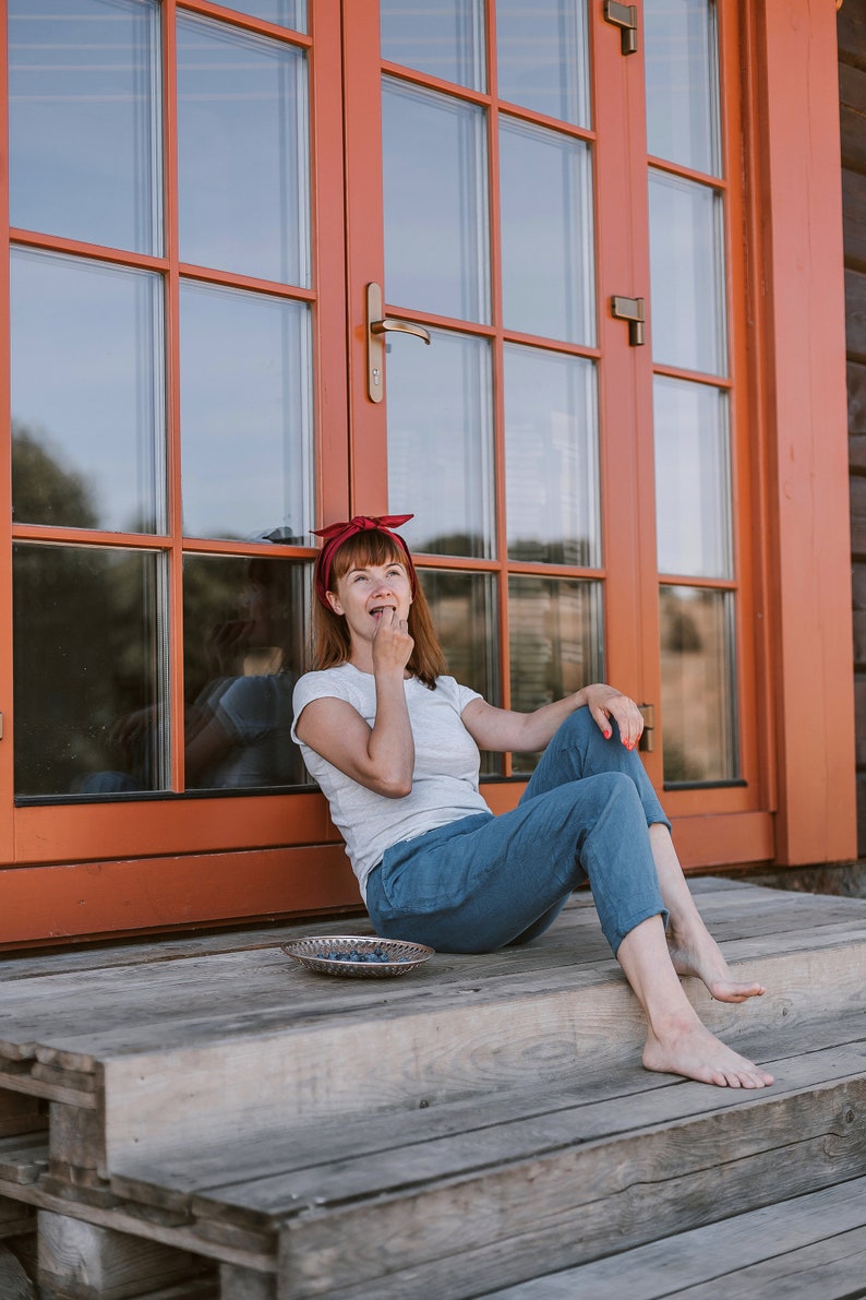 May include: A woman wearing a red headband sits on a wooden porch with a bowl of blueberries in front of her. She is eating a blueberry and looking up.