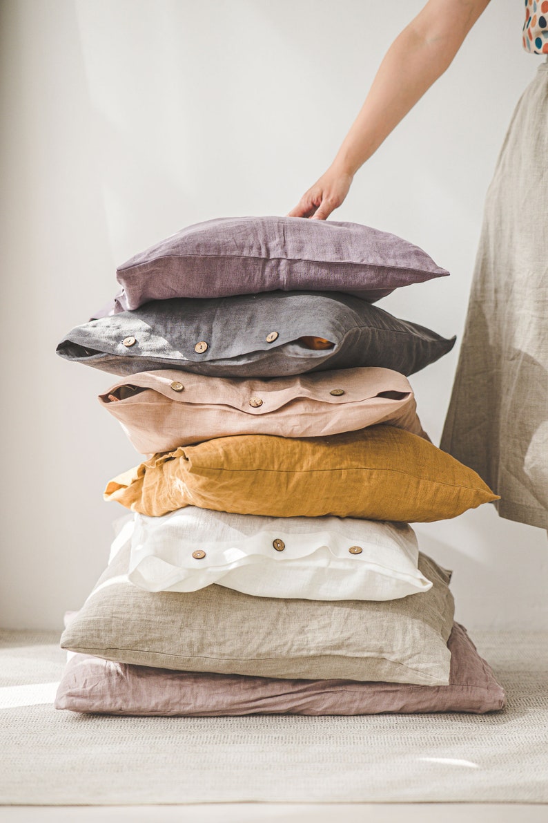May include: A stack of six linen pillow covers in various colors, including purple, gray, pink, yellow, white, and beige. The pillow covers have wooden buttons and are arranged in a pyramid shape.