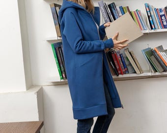 May include: A long, hooded blue coat with a full-length zipper. The coat is paired with dark blue jeans and brown slip-on shoes. The background features a bookshelf filled with books.