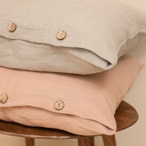May include: Two linen pillowcases, one in a light beige color and one in a light peach color, stacked on top of each other on a wooden stool. Both pillowcases have wooden buttons.