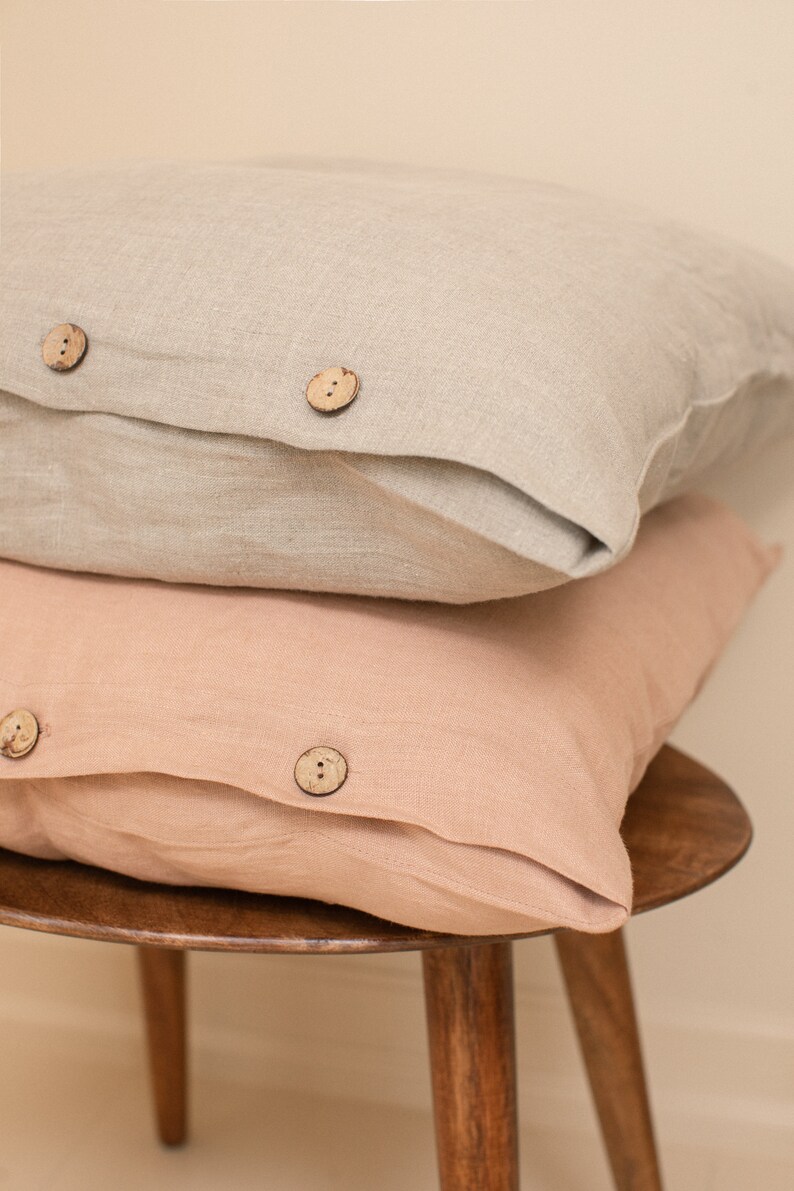 May include: Two linen pillowcases, one in a light beige color and one in a light peach color, stacked on top of each other on a wooden stool. Both pillowcases have wooden buttons.