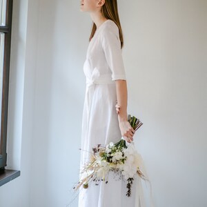 White linen wrap wedding dress with three quarter sleeves, bride holding a bouquet